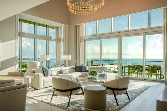 Luxury living room with cream sofas, glass coffee table, floor-to-ceiling windows and ocean views under a large chandelier.