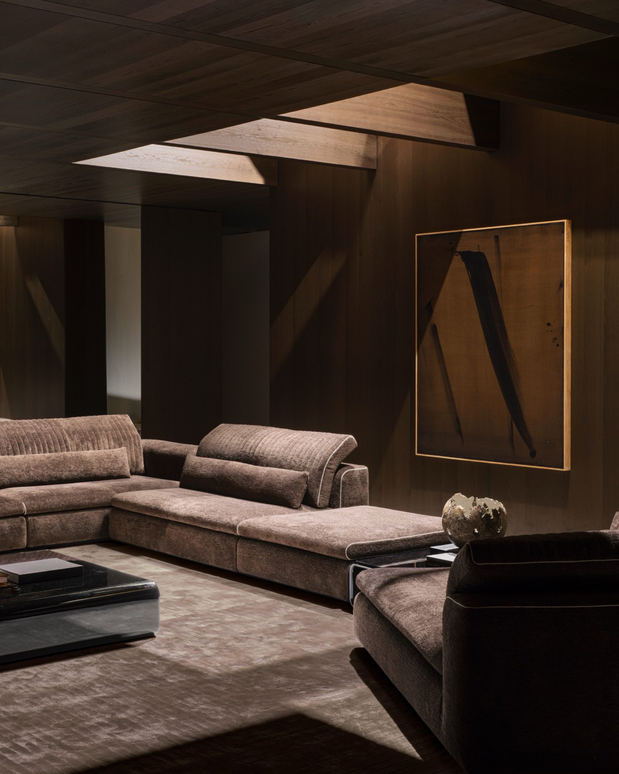 Modern living room with brown velvet sectional sofas, wood-panelled walls and ceiling, and large abstract artwork.